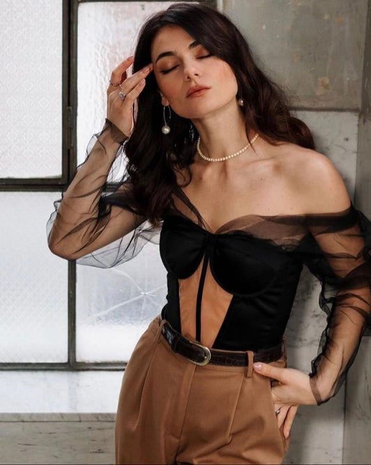 Woman wearing a black top with sheer sleeves and brown pants in a room with large windows.